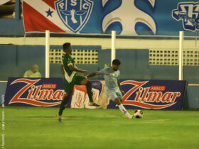 Jogador de camisa azul e cruzando a bola na área enquanto um outro jogador, de verde, tenta tirar a bola