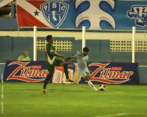 Jogador de camisa azul e cruzando a bola na área enquanto um outro jogador, de verde, tenta tirar a bola
