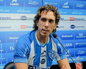 Jogador de futebol de cabelos longos vestido com camisa listrada azul e branca diante de um microfone