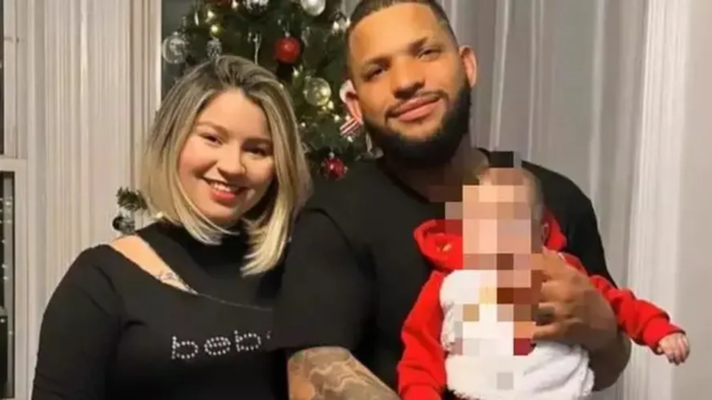 Mulher e homem de blusa branca com uma criança de roupa vermelha e branca no colo em uma casa