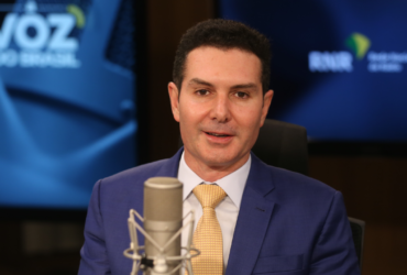 Homem com paletó azul e gravada dourada falando em um microfone