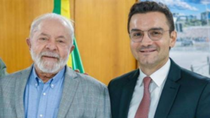 À esquerda um homem idoso, grisalho, paletó cinza e blusa azul. À direita um homem de meia idade, de paletó preto e gravata vermelha.