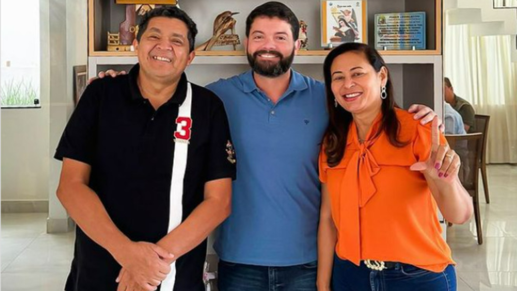 Dois homens, um de blusa preta e outro de blusa azul, e uma mulher, de laranja, sorrindo para uma fotografia