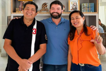 Dois homens, um de blusa preta e outro de blusa azul, e uma mulher, de laranja, sorrindo para uma fotografia