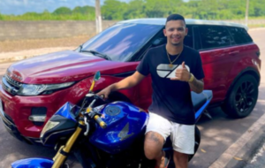 Homem posando para foto mostrando carro e moto de luxo