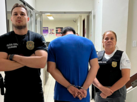 Policiais de preto e suspeito preso de azul em uma delegacia com pintura branca