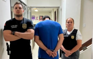 Policiais de preto e suspeito preso de azul em uma delegacia com pintura branca