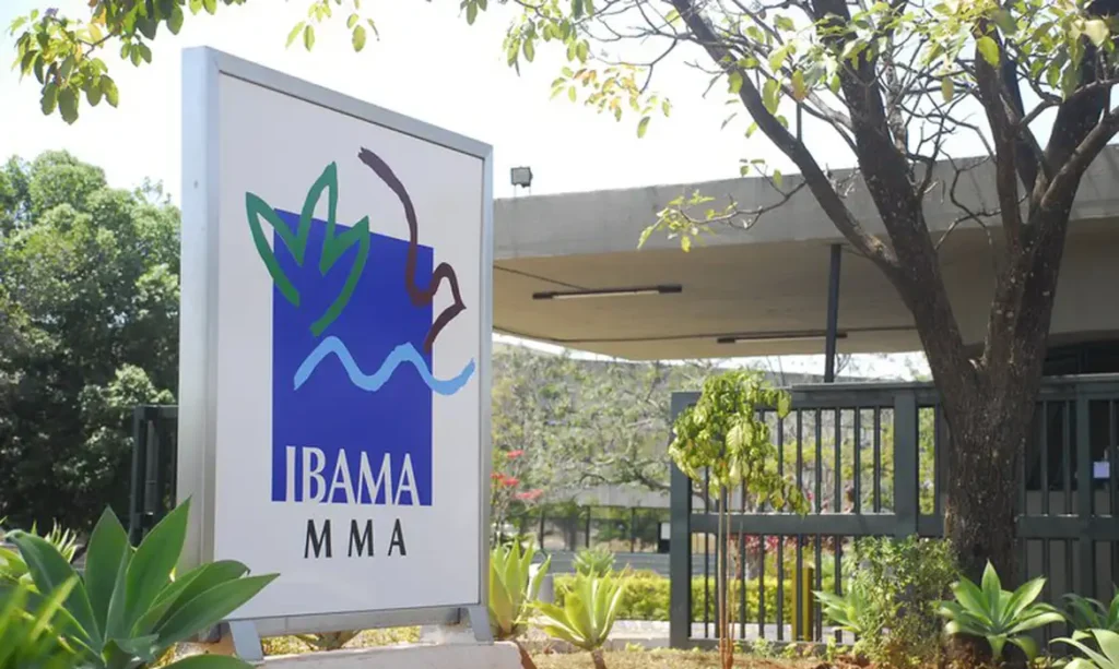 Placa do Ibama em frente à sede do órgão, com vegetação e parte do prédio ao fundo.
