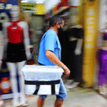 Homem caminha em uma calçada comercial carregando uma caixa plástica presa ao corpo por alça. O fundo está desfocado, com manequins e lojas ao redor, indicando movimento em área urbana.