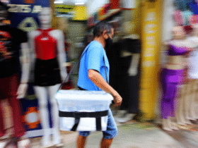 Homem caminha em uma calçada comercial carregando uma caixa plástica presa ao corpo por alça. O fundo está desfocado, com manequins e lojas ao redor, indicando movimento em área urbana.