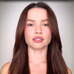 Imagem mostra uma mulher jovem olhando diretamente para a câmera, com expressão séria e neutra. Ela está em fundo claro e aparece do peito para cima.