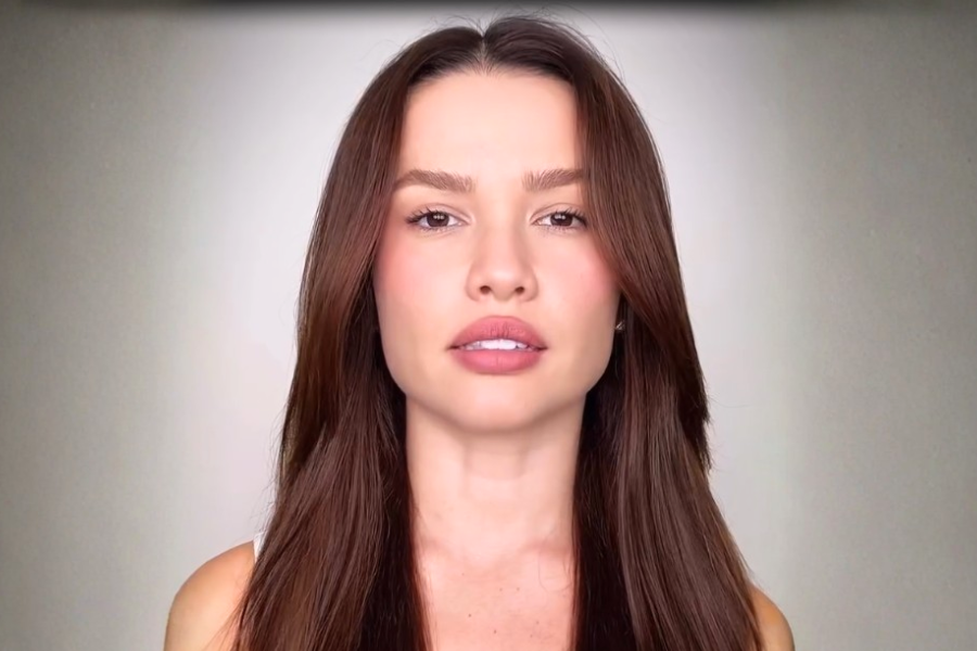 Imagem mostra uma mulher jovem olhando diretamente para a câmera, com expressão séria e neutra. Ela está em fundo claro e aparece do peito para cima.