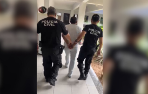Dois agentes da polícia conduzem um homem detido, segurando-o pelos braços.