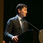 Prefeito de Minneapolis, Jacob Frey, sorrindo sutilmente em um pulpito