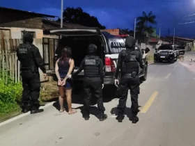 Mulher com as mãos algemadas é conduzida por quatro policiais civis em uma rua residencial durante operação policial; viaturas estão estacionadas ao fundo.