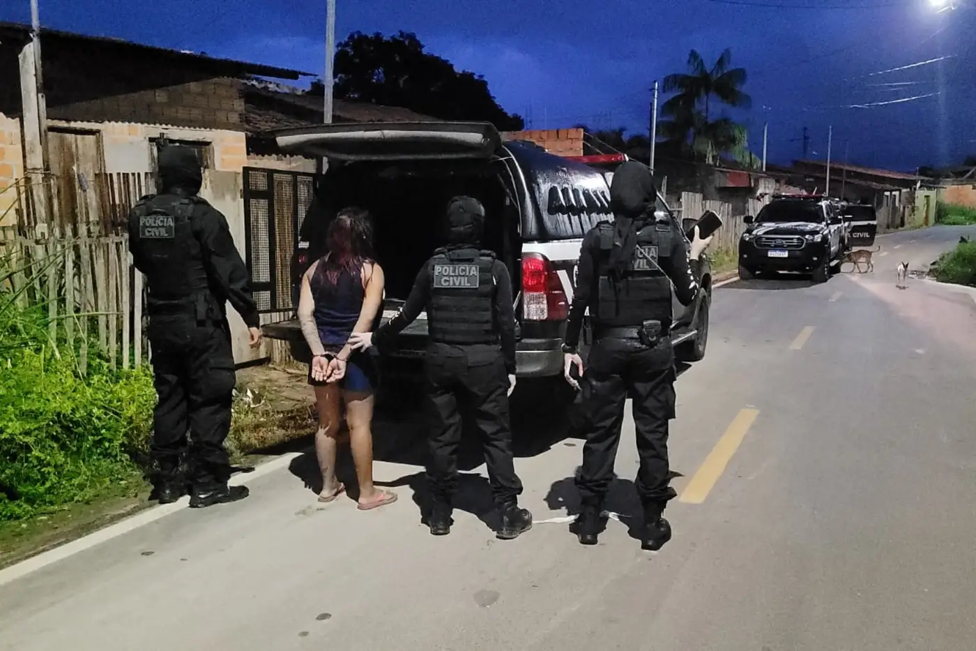 Mulher com as mãos algemadas é conduzida por quatro policiais civis em uma rua residencial durante operação policial; viaturas estão estacionadas ao fundo.