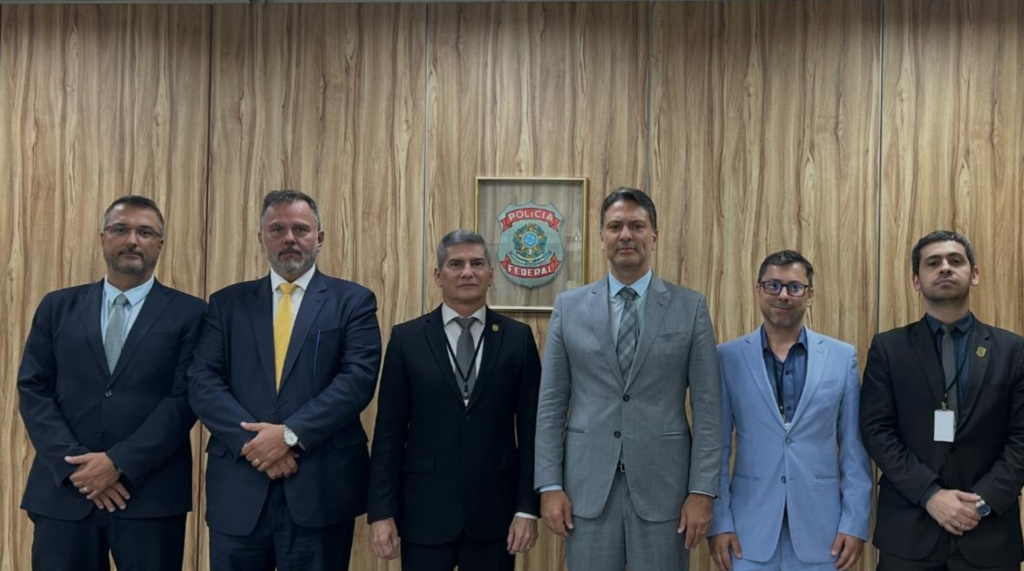 Autoridades da Polícia Civil e da Polícia Federal durante assinatura de acordo de cooperação, reunidas em sala com mesa e bandeiras ao fundo.