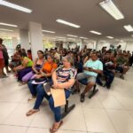 Dezenas de pessoas sentadas em sala de atendimento, aguardando com documentos em mãos.