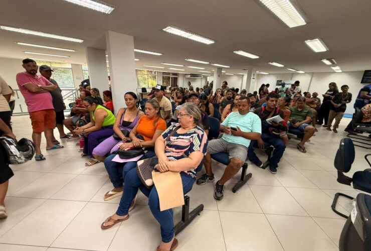Dezenas de pessoas sentadas em sala de atendimento, aguardando com documentos em mãos.
