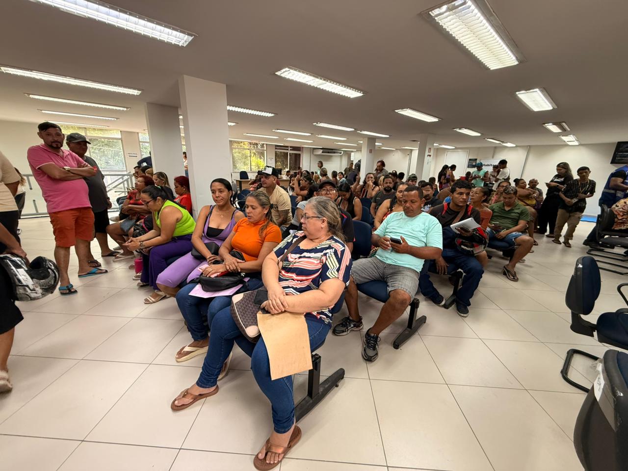 Dezenas de pessoas sentadas em sala de atendimento, aguardando com documentos em mãos.