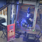 Imagem de câmera de segurança de uma conveniência onde um homem está caído na calçada do estabelecimento enquanto o outro homem que está de pé usando capacete aponta um revólver para ele.