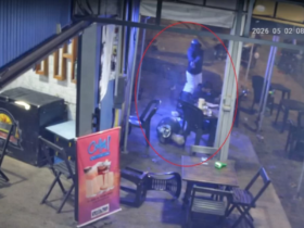 Imagem de câmera de segurança de uma conveniência onde um homem está caído na calçada do estabelecimento enquanto o outro homem que está de pé usando capacete aponta um revólver para ele.