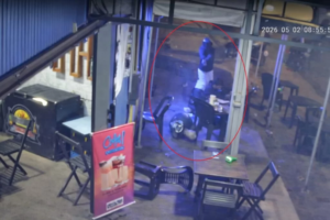 Imagem de câmera de segurança de uma conveniência onde um homem está caído na calçada do estabelecimento enquanto o outro homem que está de pé usando capacete aponta um revólver para ele.