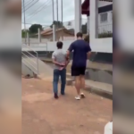 Imagem de um agente da Polícia Civil conduzindo o suspeito algemado na entrada da delegacia.