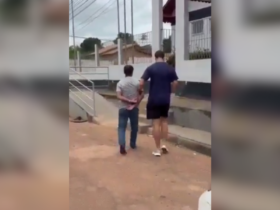 Imagem de um agente da Polícia Civil conduzindo o suspeito algemado na entrada da delegacia.