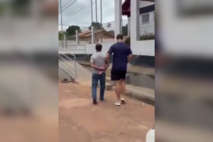 Imagem de um agente da Polícia Civil conduzindo o suspeito algemado na entrada da delegacia.