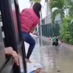 A imagem registra um ônibus parado no meio de uma rua alagada e uma passageira vestindo blusa rosa e calça jeans com metade do corpo para fora da janela enquanto tenta sair.