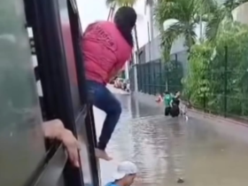 A imagem registra um ônibus parado no meio de uma rua alagada e uma passageira vestindo blusa rosa e calça jeans com metade do corpo para fora da janela enquanto tenta sair.