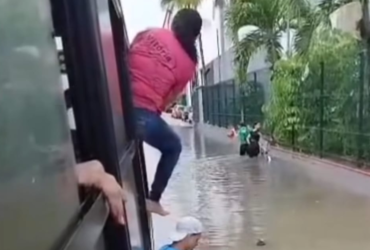 A imagem registra um ônibus parado no meio de uma rua alagada e uma passageira vestindo blusa rosa e calça jeans com metade do corpo para fora da janela enquanto tenta sair.