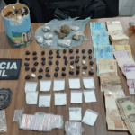 Na imagem aparece uma mesa onde estão todas as apreensões realizadas, com papelotes de substância análoga ao crack, nove papelotes maiores de substância semelhante à maconha e outros 12 papelotes de material análogo à cocaína. Além disso, tem dinheiro em espécie, uma lata cheia de moedas e os selos da Polícia Civil.