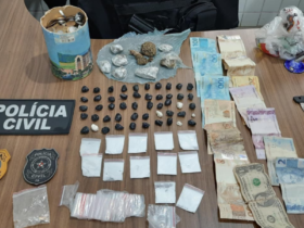 Na imagem aparece uma mesa onde estão todas as apreensões realizadas, com papelotes de substância análoga ao crack, nove papelotes maiores de substância semelhante à maconha e outros 12 papelotes de material análogo à cocaína. Além disso, tem dinheiro em espécie, uma lata cheia de moedas e os selos da Polícia Civil.