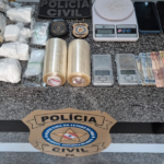 Na imagem aparece as apreensões sobre uma mesa de mármore, nela estão balaças de precisão, quantia em dinheiro, 2 rolos de plástico filme, drogas, além do selo da polícia civil.