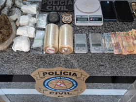 Na imagem aparece as apreensões sobre uma mesa de mármore, nela estão balaças de precisão, quantia em dinheiro, 2 rolos de plástico filme, drogas, além do selo da polícia civil.