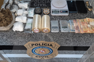 Na imagem aparece as apreensões sobre uma mesa de mármore, nela estão balaças de precisão, quantia em dinheiro, 2 rolos de plástico filme, drogas, além do selo da polícia civil.