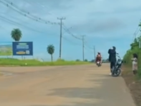 A imagem é um recorte do vídeo onde um homem aparece se deslocando com uma criança em uma motocicleta em uma estrada.