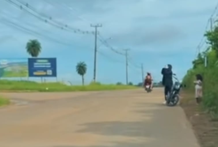 A imagem é um recorte do vídeo onde um homem aparece se deslocando com uma criança em uma motocicleta em uma estrada.