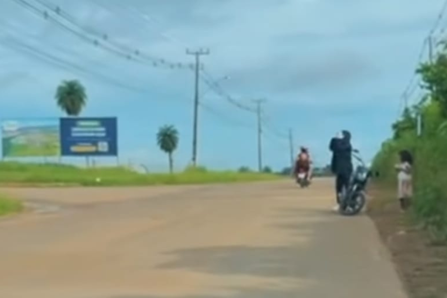 A imagem é um recorte do vídeo onde um homem aparece se deslocando com uma criança em uma motocicleta em uma estrada.
