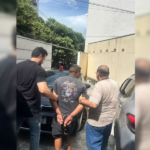 Na imagem o suspeito aparece de costas algemado, enquanto dois agentes da Polícia Civil sem farda o conduzem em meio a um estacionamento com veículos.