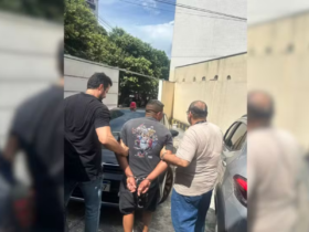 Na imagem o suspeito aparece de costas algemado, enquanto dois agentes da Polícia Civil sem farda o conduzem em meio a um estacionamento com veículos.