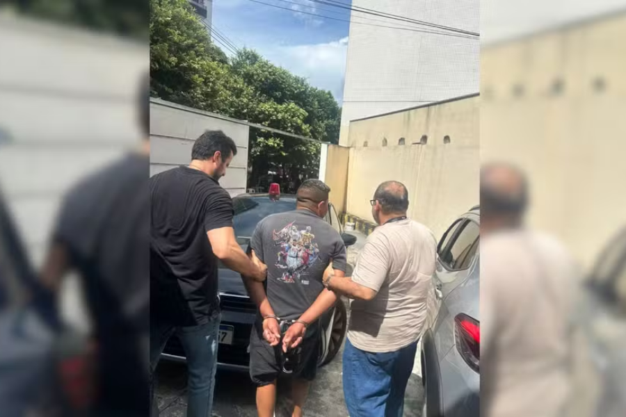 Na imagem o suspeito aparece de costas algemado, enquanto dois agentes da Polícia Civil sem farda o conduzem em meio a um estacionamento com veículos.