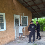Na imagem aparece a frente de uma casa amarela e quatro agentes fardados da polícia civil na entrada, junto de um suspeito.