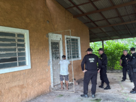 Na imagem aparece a frente de uma casa amarela e quatro agentes fardados da polícia civil na entrada, junto de um suspeito.