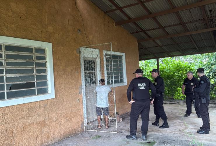 Na imagem aparece a frente de uma casa amarela e quatro agentes fardados da polícia civil na entrada, junto de um suspeito.
