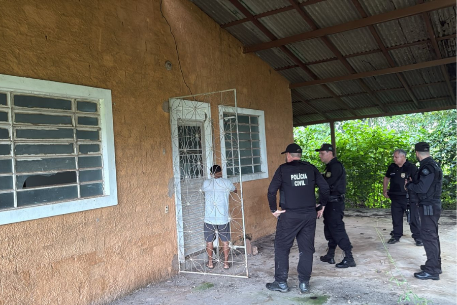 Na imagem aparece a frente de uma casa amarela e quatro agentes fardados da polícia civil na entrada, junto de um suspeito.