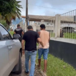 Imagem do suspeito sendo coduzido por dois agentes até a delegacia após a prisão, ao lado de uma caminhonete cinza.