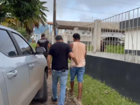 Imagem do suspeito sendo coduzido por dois agentes até a delegacia após a prisão, ao lado de uma caminhonete cinza.